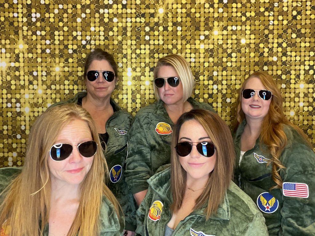 A trivia team in matching flight suits and aviator sunglasses posing in front of InstaGlee's gold sequin backdrop